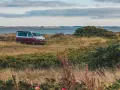 Husbil parkerad vid en gräsbevuxen kustlinje med buskar och blommor. Vattnet glittrar i bakgrunden, en idyllisk vy över norra Danmark.