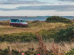 Husbil parkerad vid en gräsbevuxen kustlinje med buskar och blommor. Vattnet glittrar i bakgrunden, en idyllisk vy över norra Danmark.