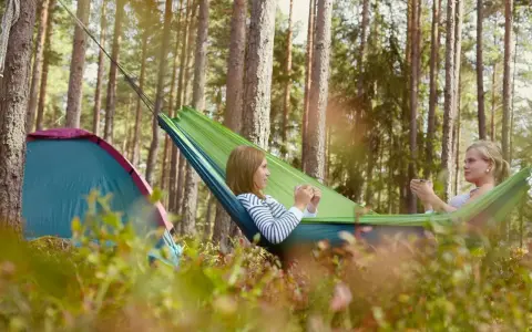 Två tjejer njuter av en lugn stund i en hängmatta på en av Sveriges fina campingar. Bredvid dem står ett tält.