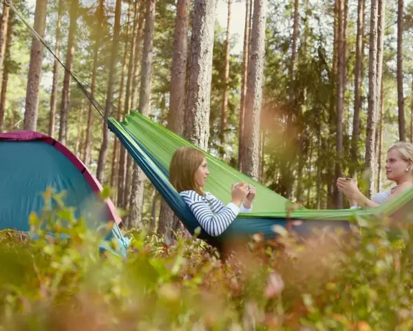 Två tjejer njuter av en lugn stund i en hängmatta på en av Sveriges fina campingar. Bredvid dem står ett tält.