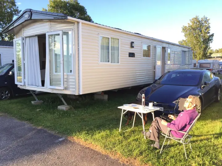 En person njuter av solen vid ett campingbord utanför en husvagn. En Tesla parkerad bredvid. Camping Timrå.