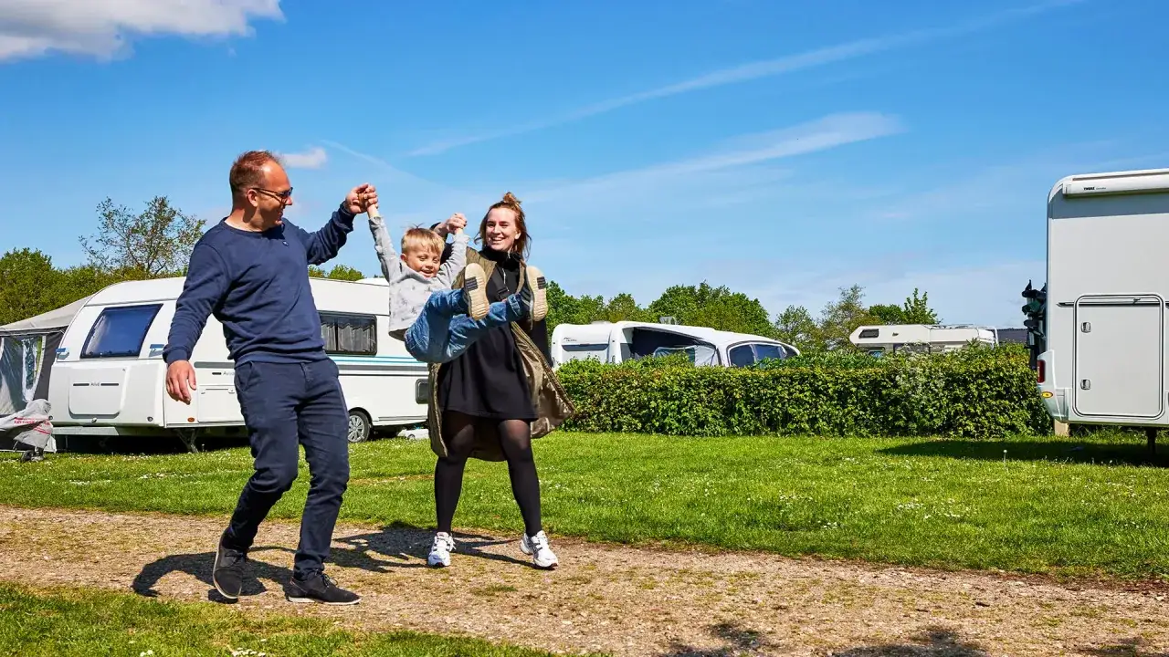 Familj njuter av en solig dag p&aring; camping i Billund. Pappa lyfter barnet medan mamman skrattar. Husvagnar och husbilar syns i bakgrunden.