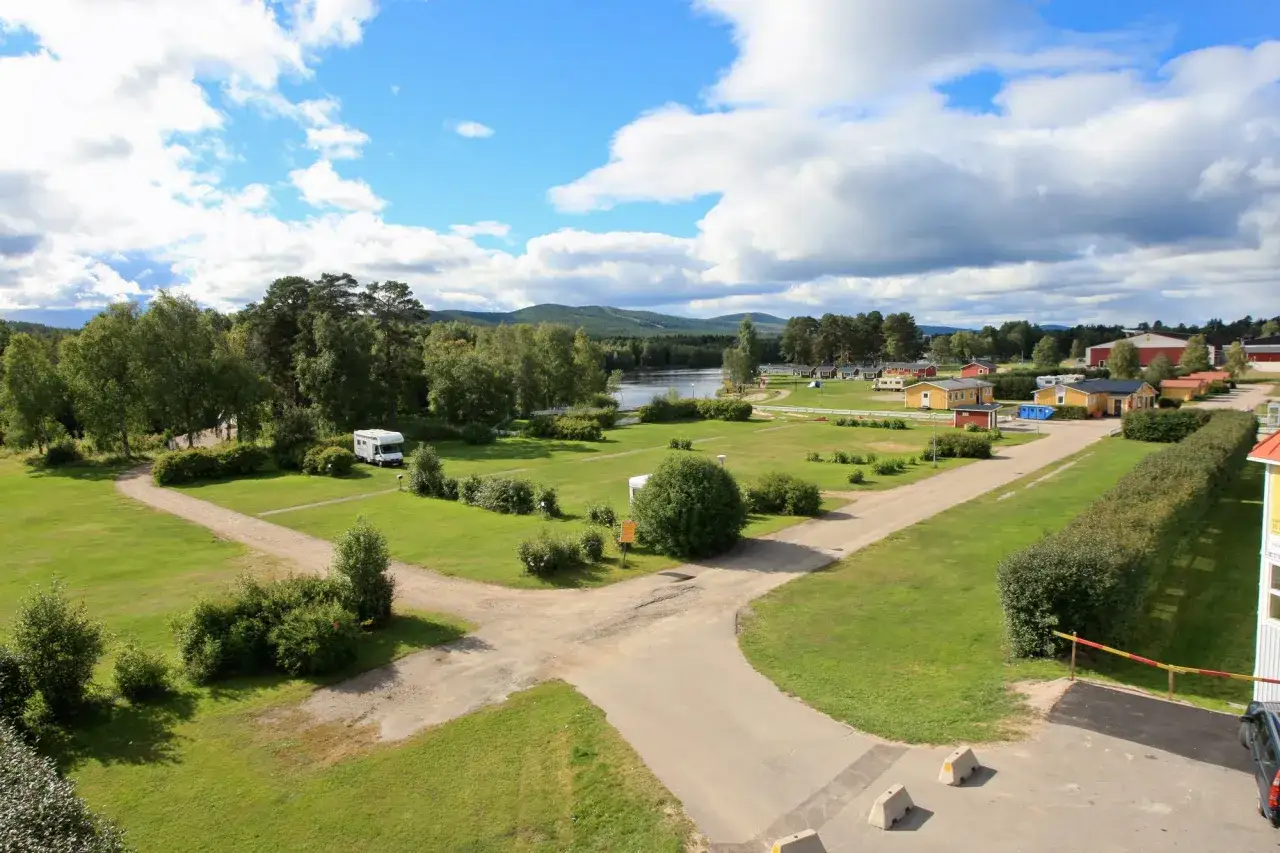 Vacker vy &ouml;ver svegs camping med husbilar, stugor och en sj&ouml; i bakgrunden under en molnig men solig dag.