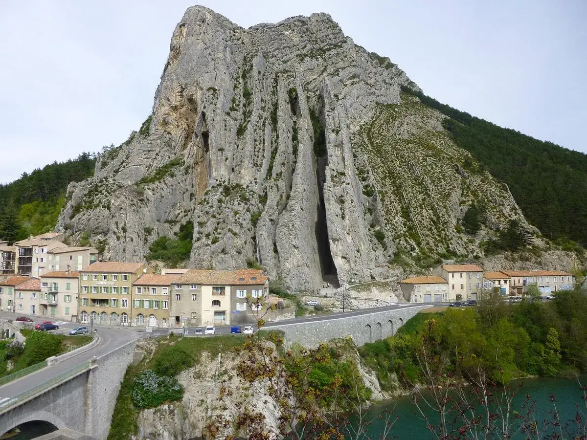 Imponerande klippformation vid Route Napoléon. En by klättrar uppför bergssluttningen, med en bro och en turkos flod nedanför.