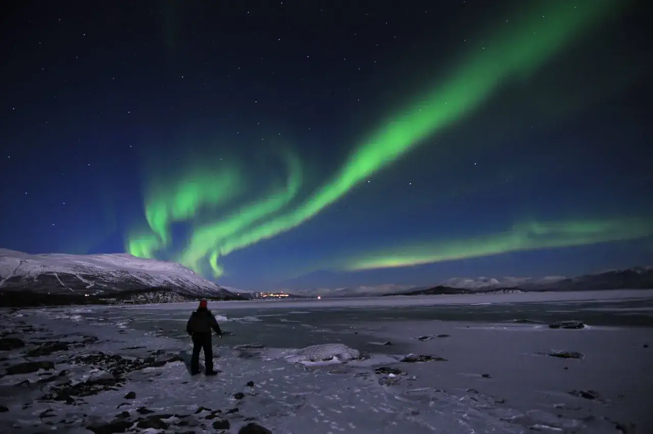 En person st&aring;r p&aring; en isig strand och ser norrsken dansa &ouml;ver en sn&ouml;kl&auml;dd bergskedja.