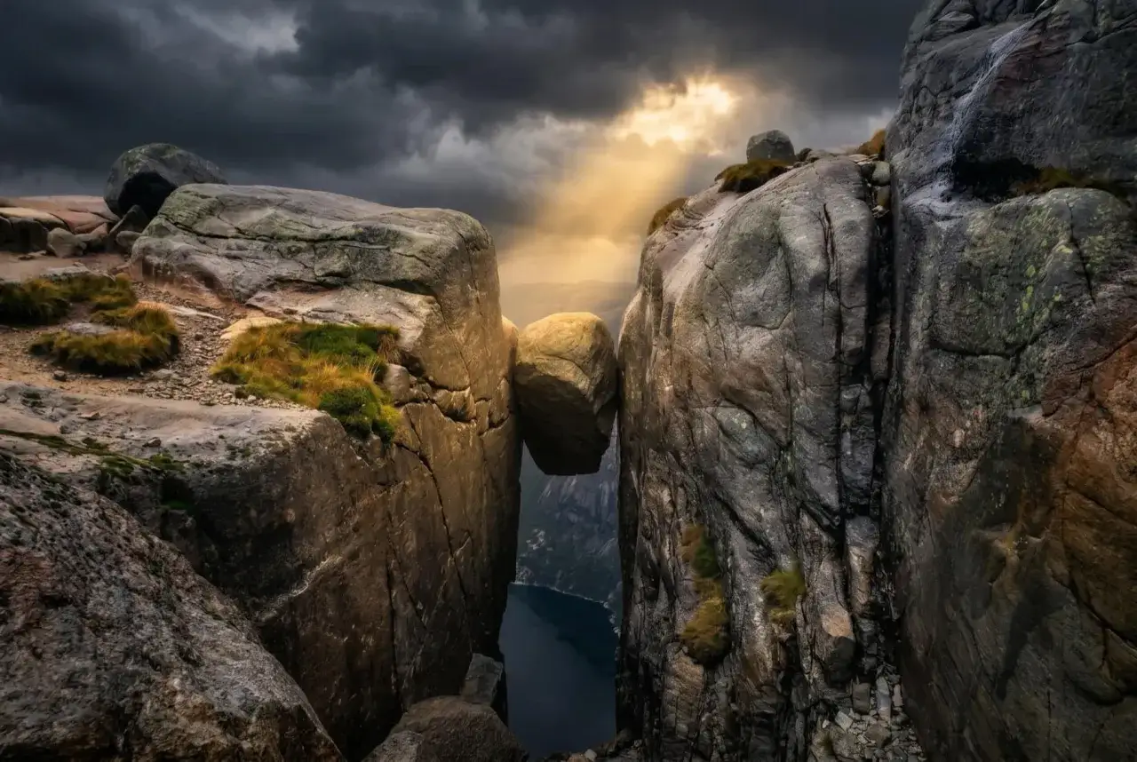 Enorm sten balanserar mellan klippor i Norge. Dramatiskt ljus bryter igenom molnen över en djup fjord.