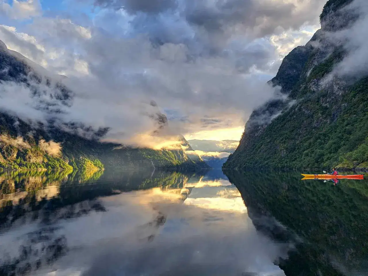 En kajakpaddlare njuter av de fina platserna i Norge, omgiven av dramatiska fjordar och moln som speglas i det lugna vattnet.