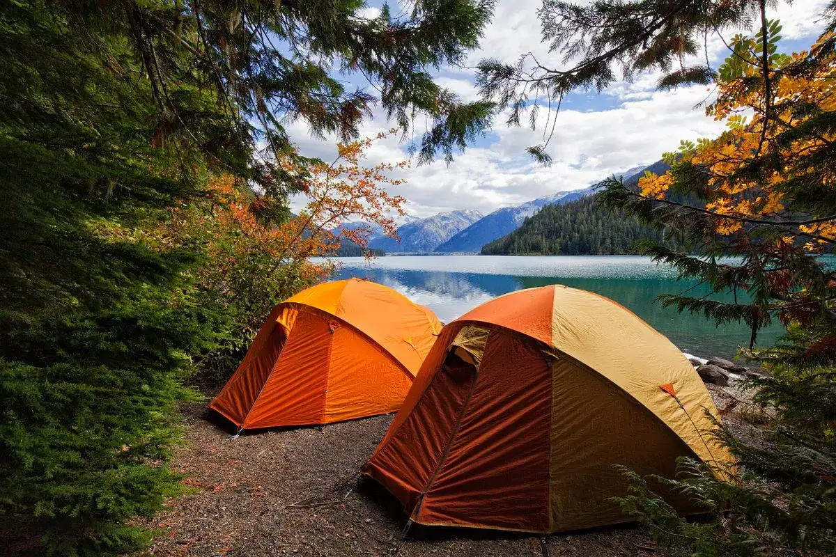 Två orange tält står uppställda vid en sjö, omgivna av höstfärgad skog och berg. En perfekt plats för kamlunge camping.