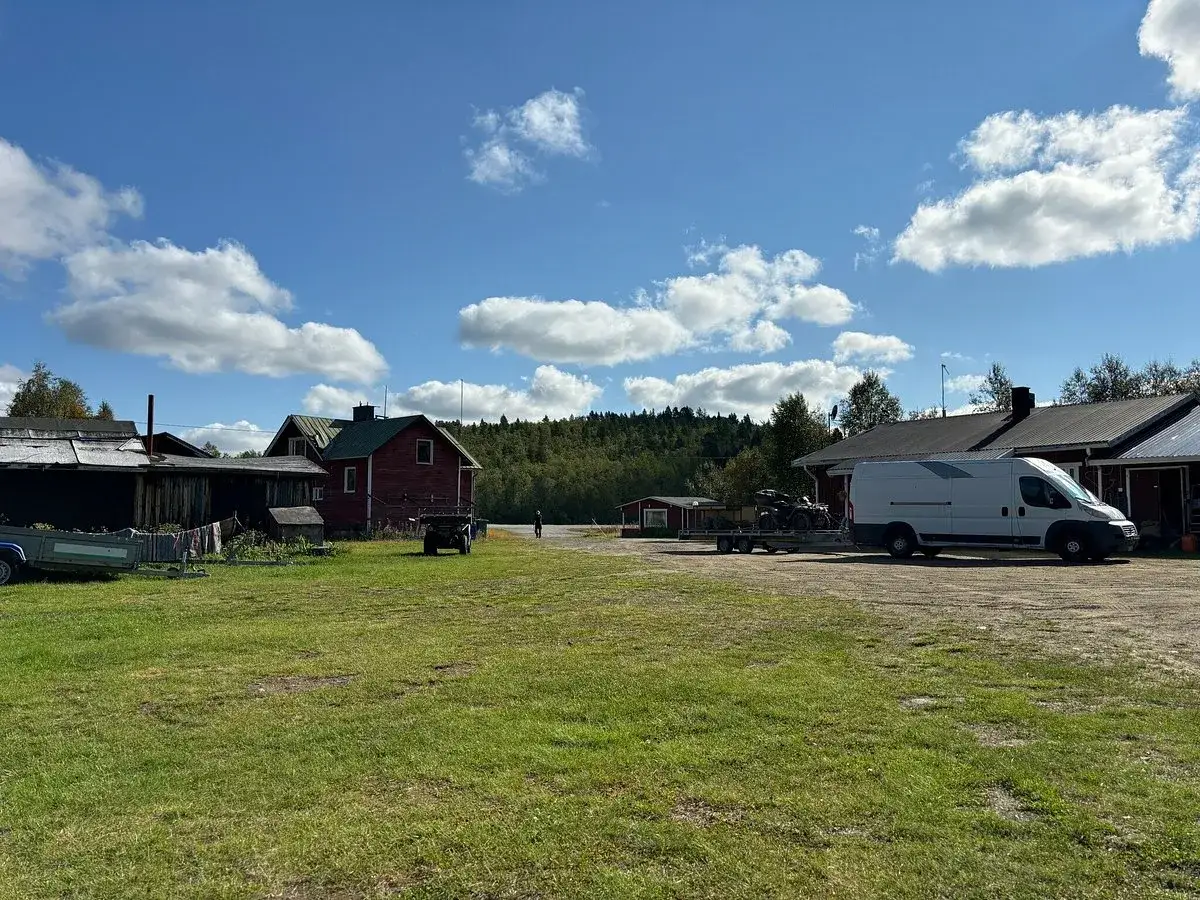 Karesuando camping: R&ouml;d stuga, vit sk&aring;pbil med sl&auml;p och fyrhjuling p&aring; gr&auml;splan under molnig himmel.
