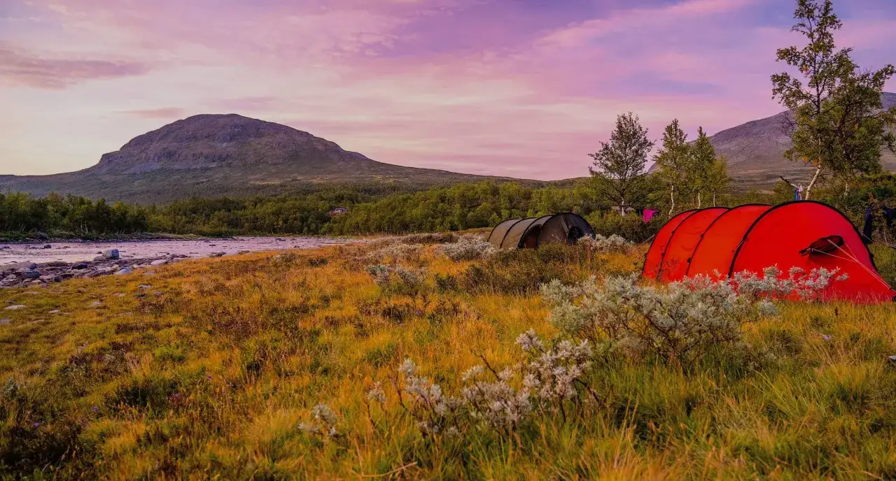 Tv&aring; t&auml;lt st&aring;r uppe i ett f&auml;lt med gulnande gr&auml;s och buskar, en flod och ett berg i bakgrunden under en rosa himmel. Perfekt f&ouml;r camping i Blekinge.