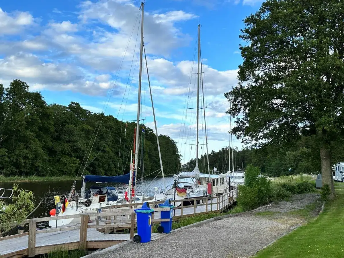 Segelb&aring;tar vid kajen p&aring; Dalbergs&aring; camping. Gr&ouml;nskande tr&auml;d och bl&aring; himmel med moln.