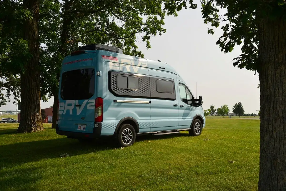 En ljusbl&aring; eldriven pl&aring;tis, en Winnebago ERV2, st&aring;r parkerad p&aring; en gr&auml;smatta under tr&auml;d.