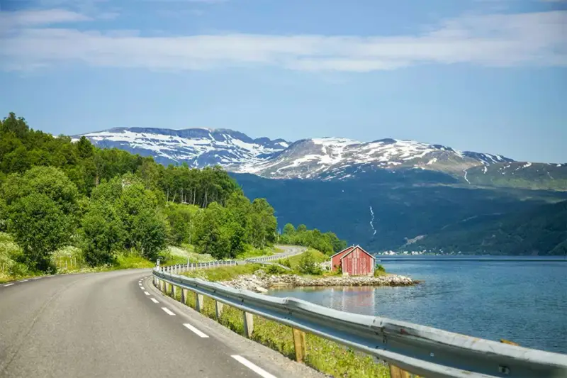 En slingrande väg längs en fjord i Norge, med snöklädda berg i bakgrunden. En röd stuga vid vattnet. Perfekt för en roadtrip norge rutt.