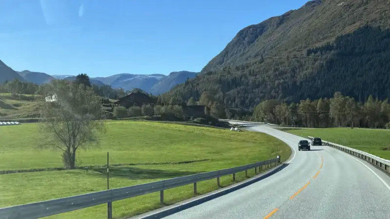 En bil kör på en slingrande väg genom ett grönt landskap med berg och hus. En perfekt start på en semester i Norge.