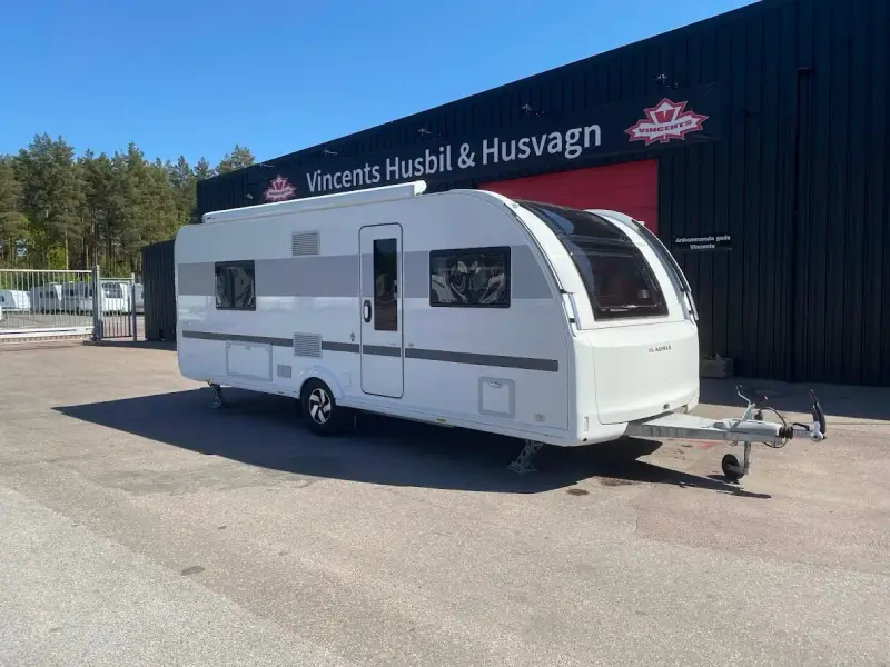 En vit Adria Alpina 613 UT husvagn står parkerad framför en byggnad med texten "Vincents Husbil & Husvagn".